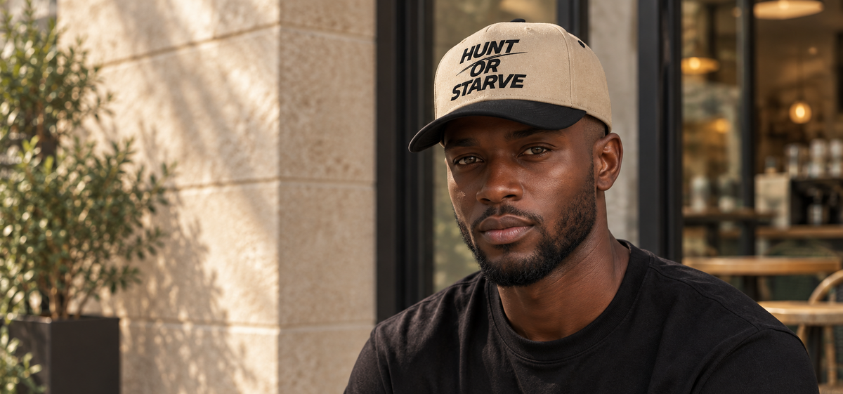A man wearing a beige and black baseball cap with the text 'HUNT OR STARVE' in black lettering, paired with a black t-shirt, photographed in an outdoor cafe setting.