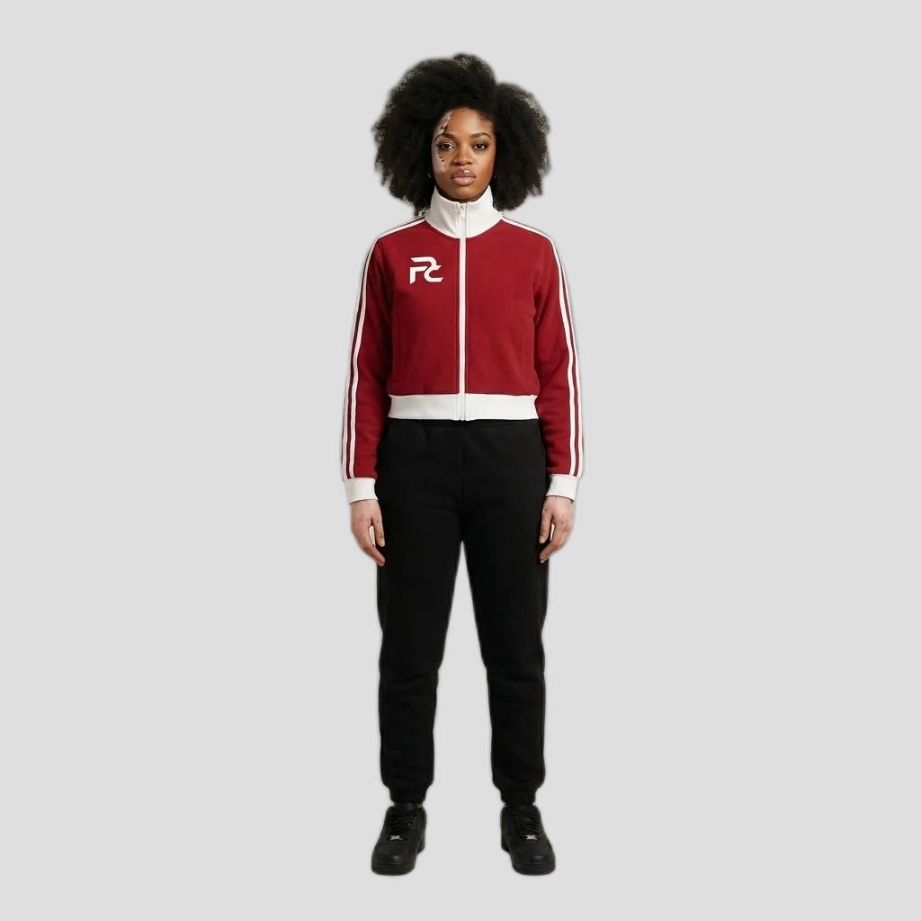 A model stands against a neutral beige studio background wearing a red and white track jacket with black sweatpants and black sneakers. The jacket features a white collar, white zipper, white side stripes, and a white 'RZ' logo on the left chest.