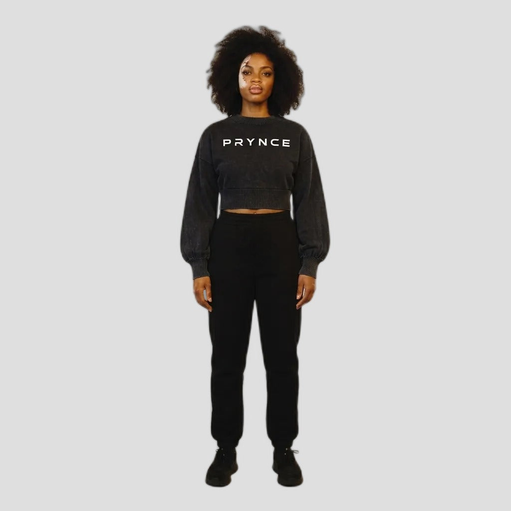 A model stands against a plain light beige studio background wearing a dark charcoal gray cropped sweatshirt with 'PRYNCE' printed in white across the chest, paired with black sweatpants and black sneakers.
