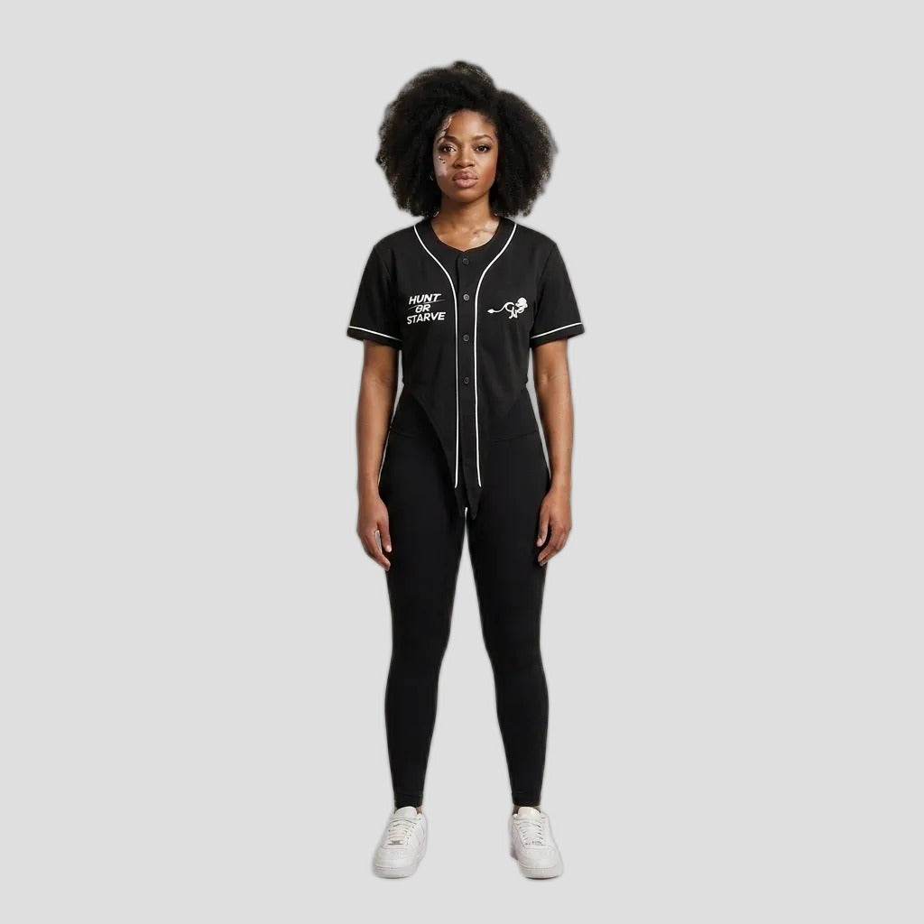 A model stands against a plain beige studio background wearing a black baseball-style jumpsuit with white piping and graphics, black leggings, and white sneakers.