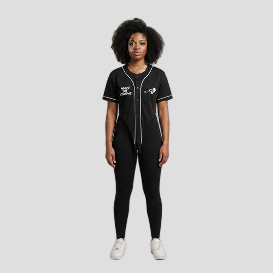 A model stands against a plain beige studio background wearing a black baseball-style jumpsuit with white piping and graphics, black leggings, and white sneakers.