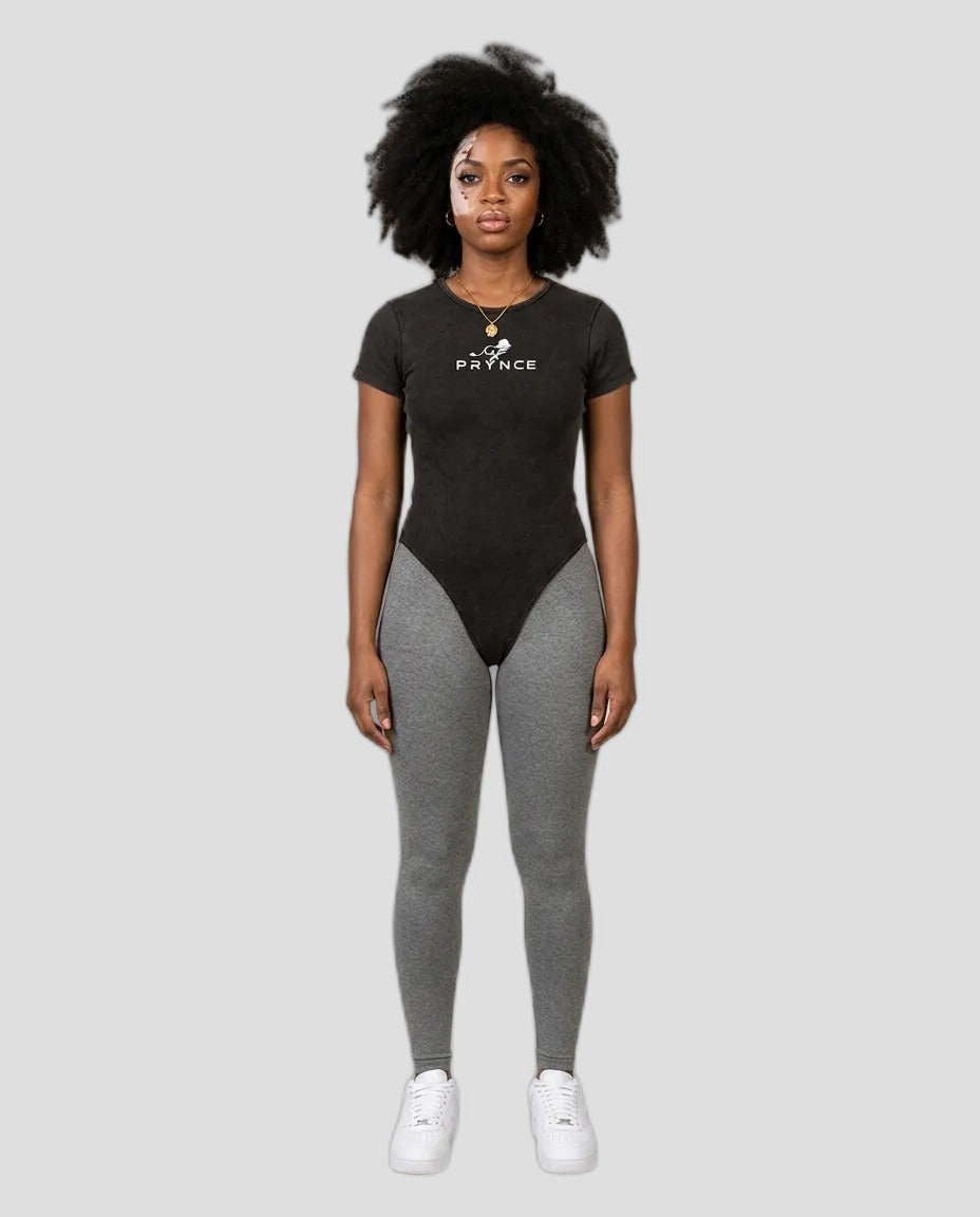 Woman wearing the PRYNCE Mineral Wash Bodysuit in black with grey leggings, studio photo
