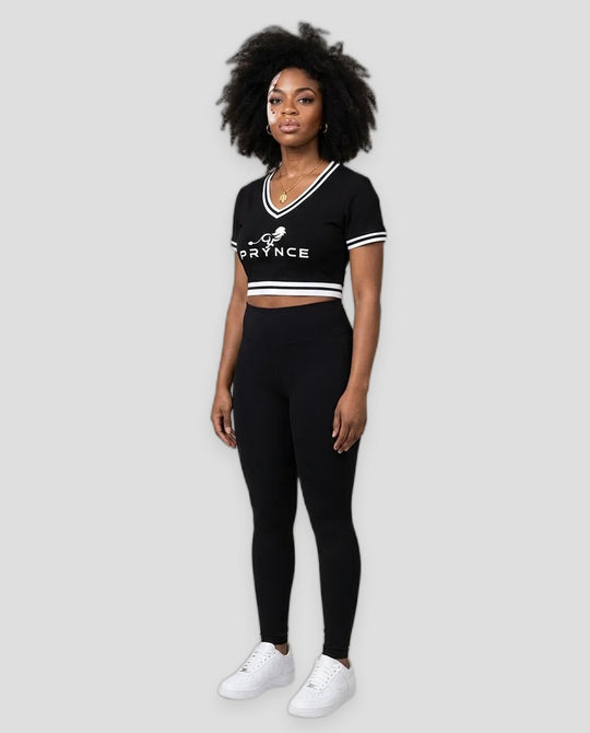 A woman modeling a black athletic crop top and matching leggings set with white trim and white sneakers, standing against a plain light beige studio background.