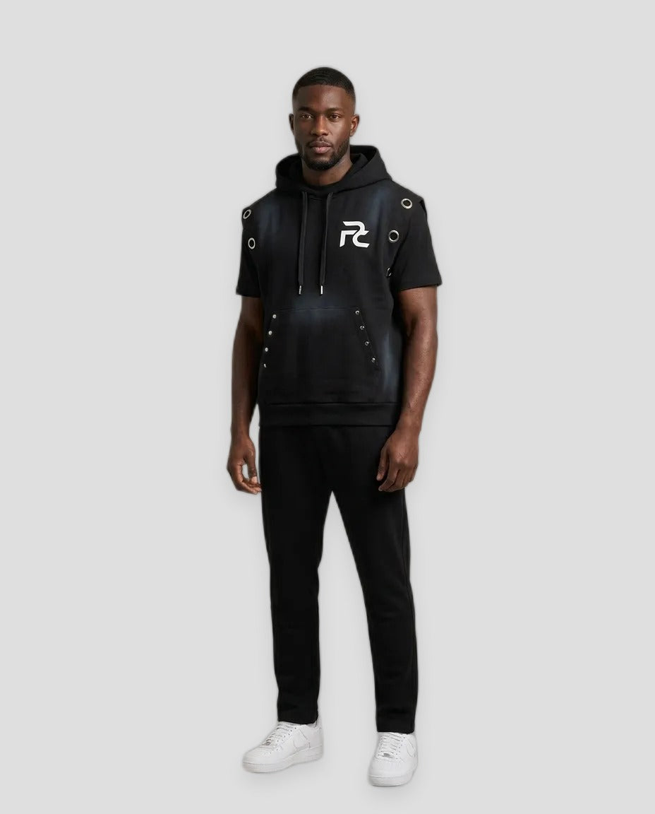 A man modeling a black short-sleeve hoodie with faux-leather paneling, silver grommet details, and a white graphic logo on the chest, paired with black sweatpants and white sneakers, against a solid beige studio background.