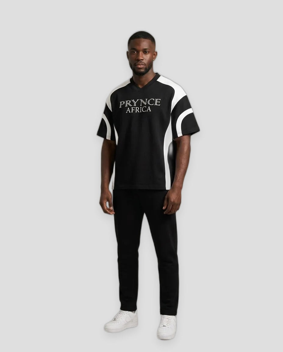 A man modeling a black short-sleeve t-shirt with white side panels and 'PRINCE AFRICA' printed in white across the chest, paired with black pants and white sneakers, against a solid beige background.