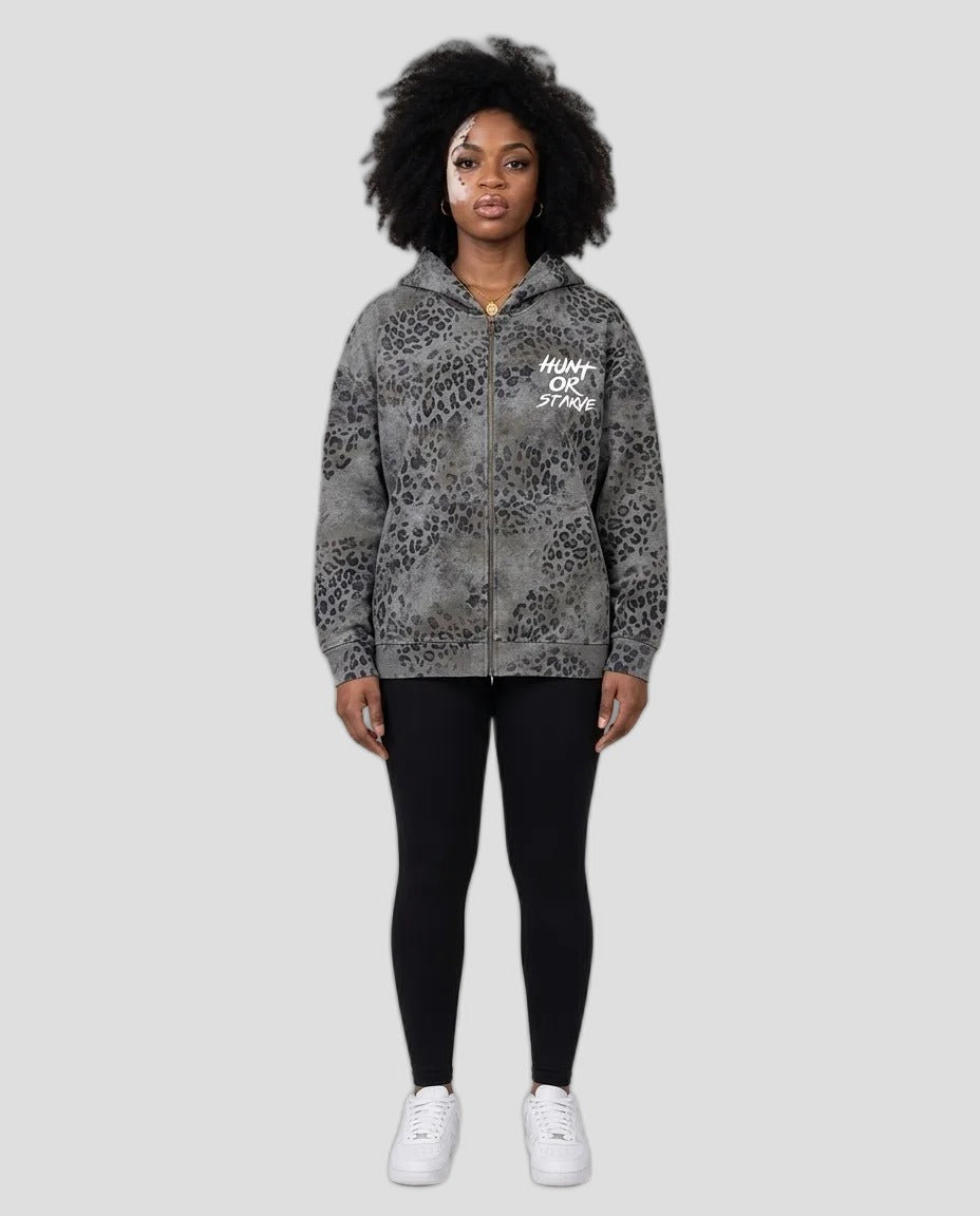 A model stands against a neutral beige studio background wearing a gray and black leopard-print zip-up hoodie with 'HUNT OR STAY' printed in white on the chest, paired with black leggings and white sneakers.