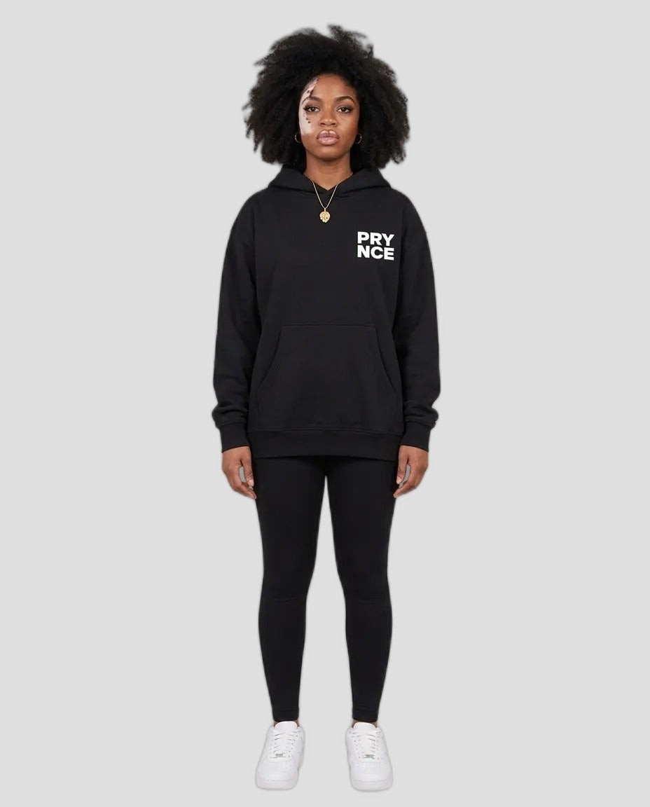 A model stands against a neutral beige studio background wearing a black pullover hoodie with 'PRY NCE' printed in white on the left chest, paired with black leggings and white sneakers.