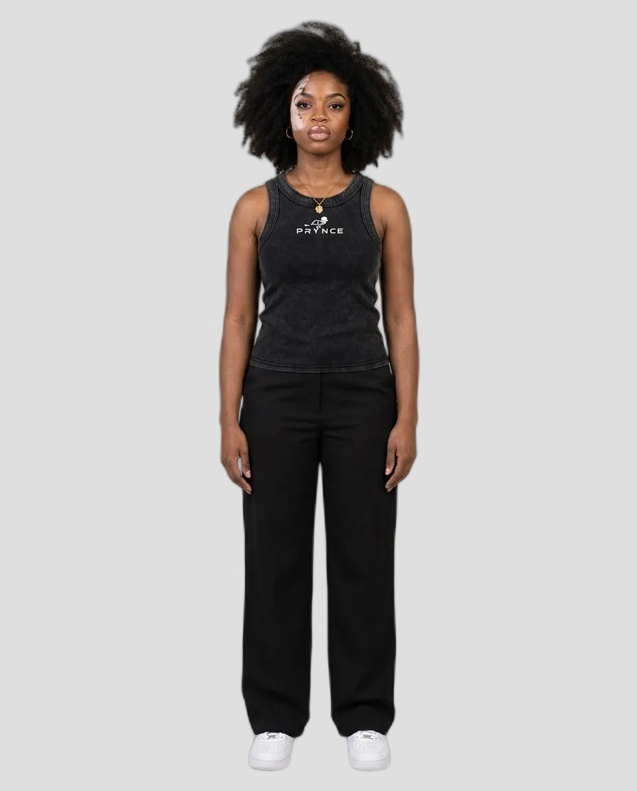 A woman stands against a plain light grey studio background wearing a dark charcoal sleeveless tank top with a white printed logo and black wide-leg pants, paired with white sneakers.
