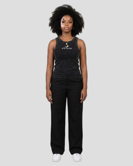 A woman stands against a plain light grey studio background wearing a dark charcoal sleeveless tank top with a white printed logo and black wide-leg pants, paired with white sneakers.