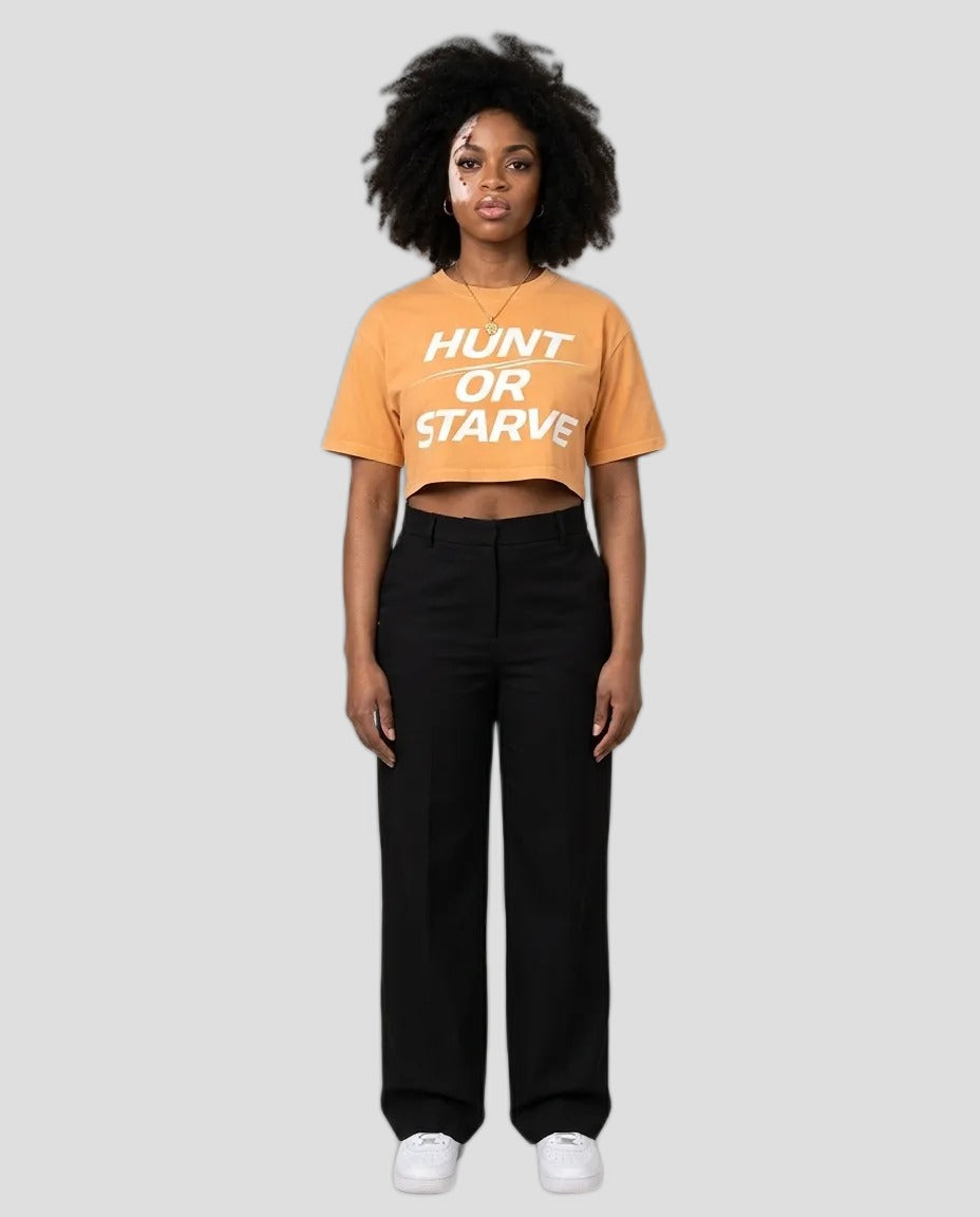 A model stands against a plain beige studio background wearing a cropped orange t-shirt with the text 'HUNT OR STARVE' in white, paired with black wide-leg pants and white sneakers.
