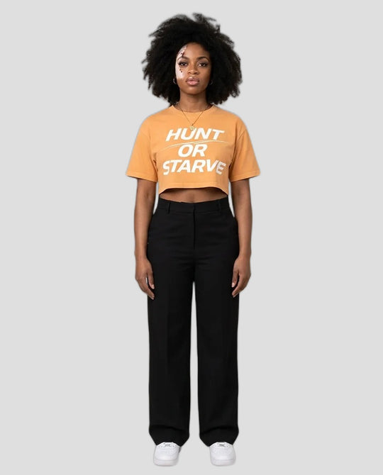 A model stands against a plain beige studio background wearing a cropped orange t-shirt with the text &