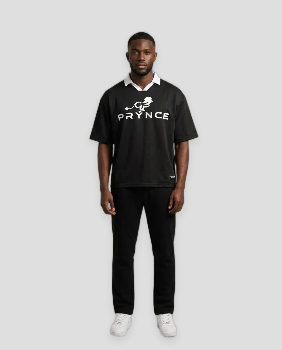 A man modeling a black short-sleeve polo shirt with a white collar, white 'PRYNCE' text and bird logo on the chest, paired with black pants and white sneakers, against a solid light beige studio background.