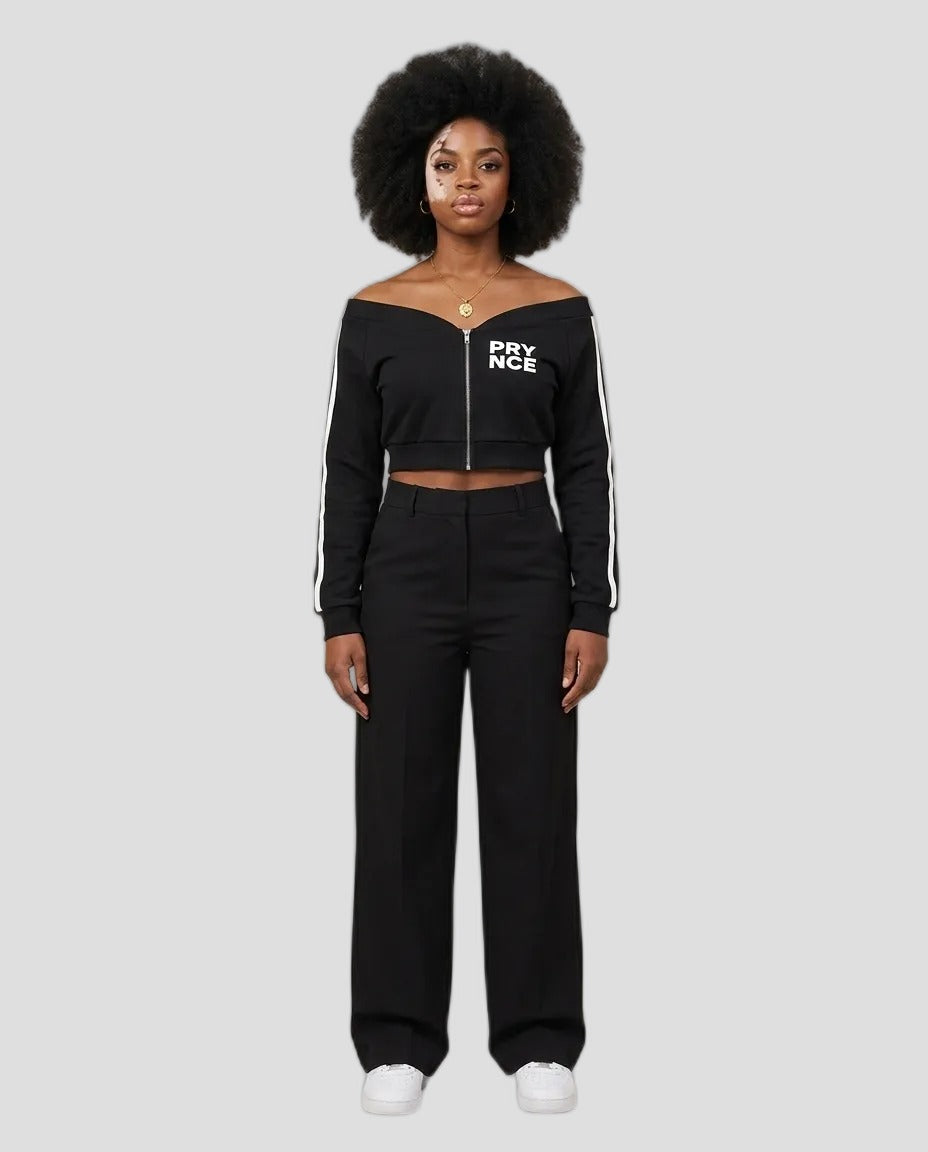 A model with an afro hairstyle stands against a neutral beige studio background wearing a black off-the-shoulder cropped zip-up top with white 'PYNCE' lettering on the chest and white side stripes, paired with black wide-leg pants and white sneakers.