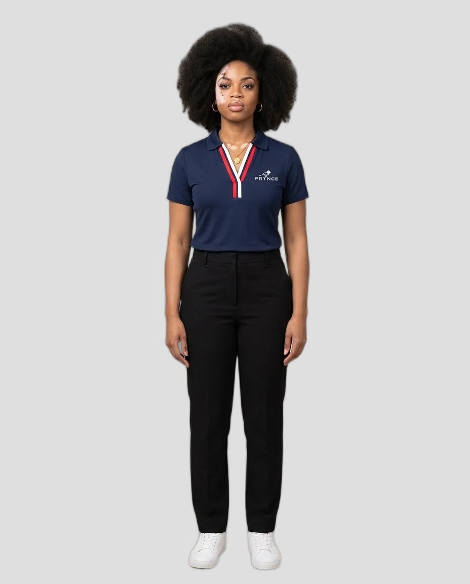 A woman stands against a plain beige studio background wearing a navy polo shirt with a red and white striped collar trim and white logo on the left chest, paired with black trousers and white sneakers.