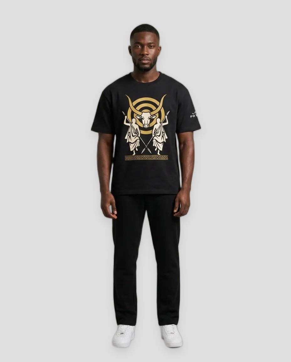 A man modeling a black short-sleeve t-shirt with a large gold and white graphic print on the front, paired with black pants and white sneakers, against a neutral light gray background.