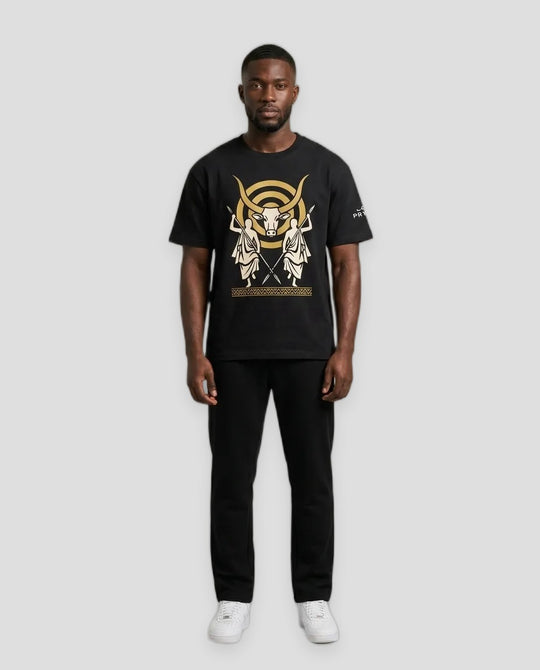 A man modeling a black short-sleeve t-shirt with a large gold and white graphic print on the front, paired with black pants and white sneakers, against a neutral light gray background.