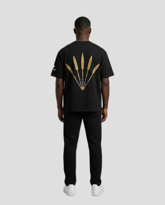 A man viewed from the back wearing a black t-shirt with a large gold graphic of three feathers on the back, paired with black pants and white sneakers, against a neutral beige background.
