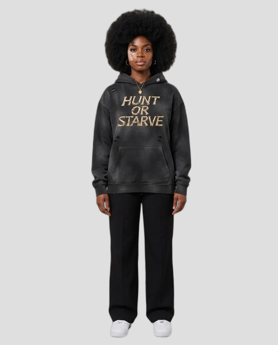 A model wearing a washed black pullover hoodie with 'HUNT OR STARVE' printed in gold across the chest, paired with black pants and white sneakers, set against a plain beige studio background.