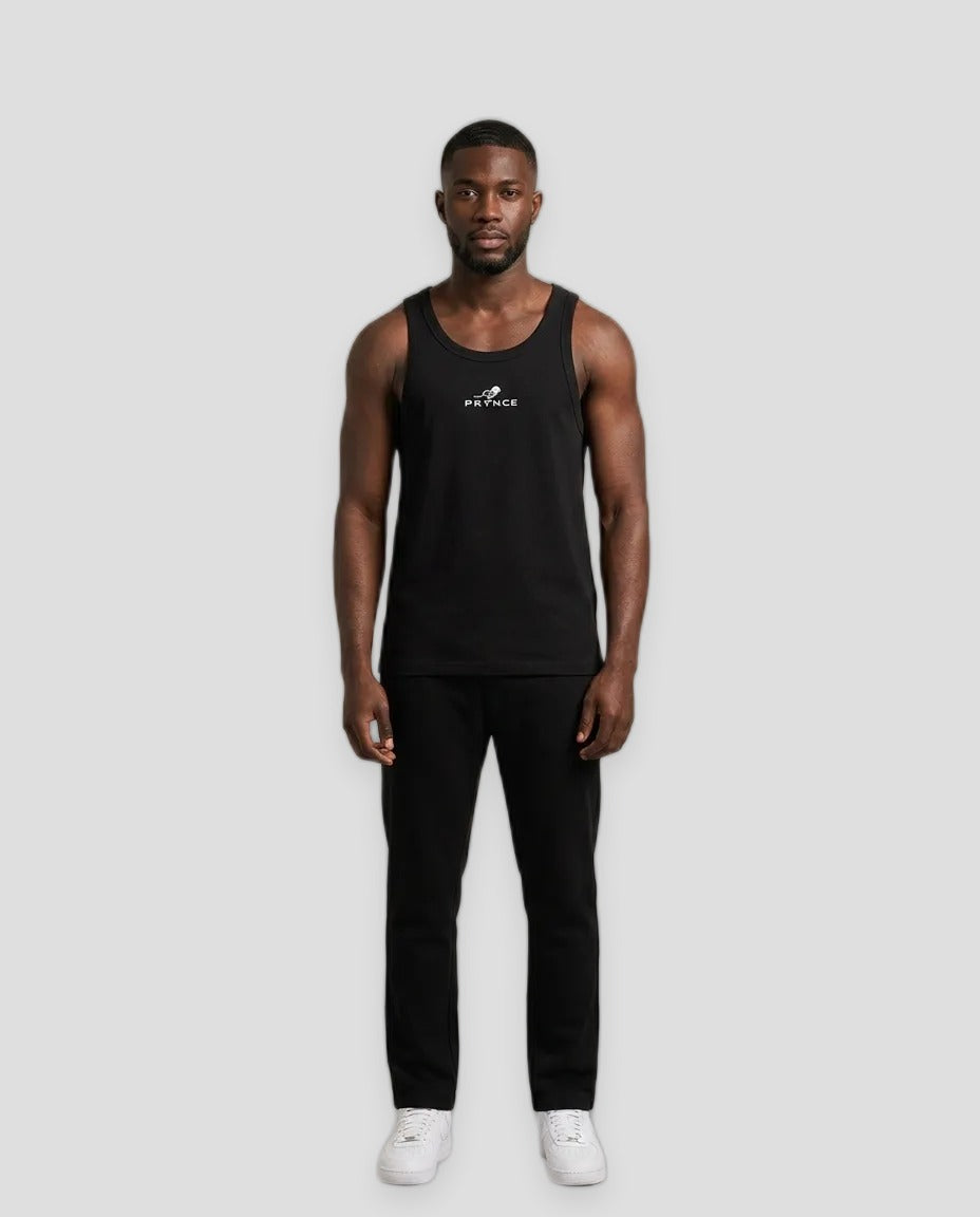 A man modeling a black sleeveless tank top with a small white logo on the chest, paired with black pants and white sneakers, standing against a plain grey studio background.