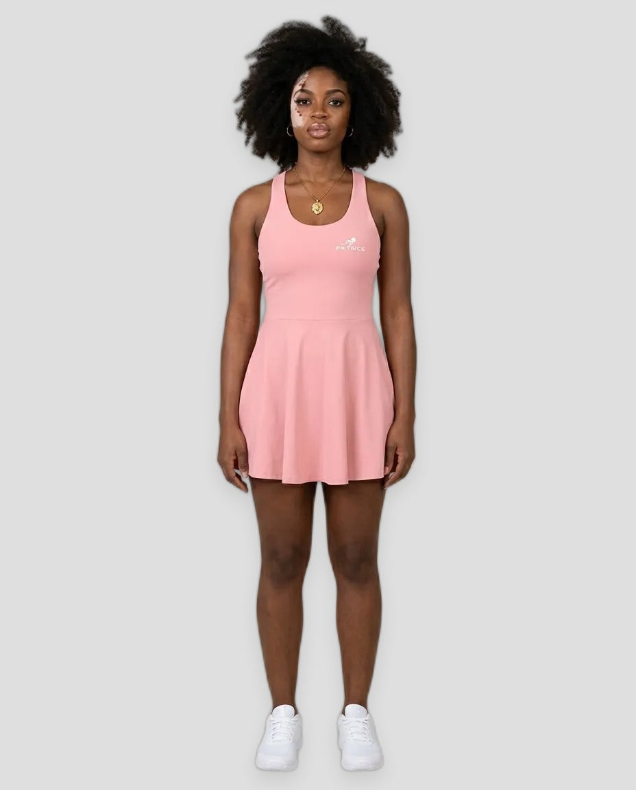 A woman modeling a light pink sleeveless athletic dress with a small white logo on the left chest, paired with white sneakers, against a solid light grey studio background.