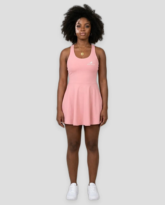 A woman modeling a light pink sleeveless athletic dress with a small white logo on the left chest, paired with white sneakers, against a solid light grey studio background.