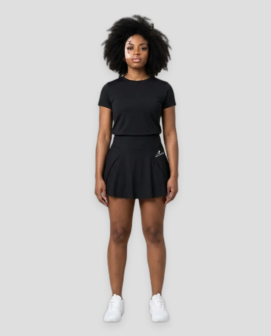 A woman modeling a black short-sleeve athletic t-shirt and matching black tennis skirt with white sneakers, standing against a light gray studio background.