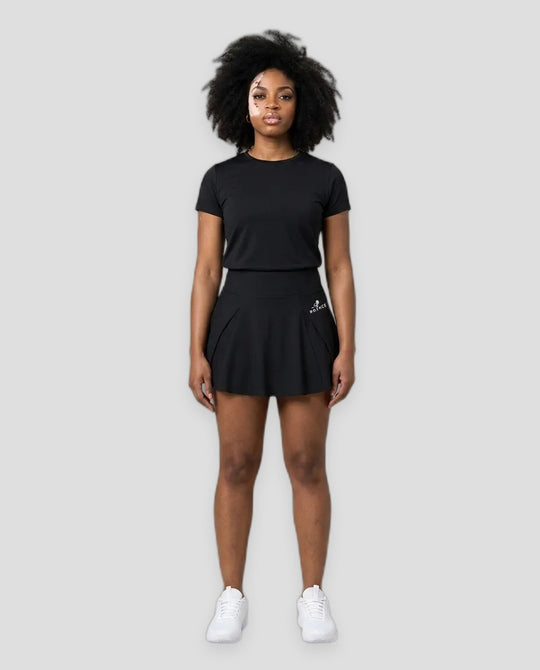 A woman modeling a black short-sleeve athletic t-shirt and matching black tennis skirt with white sneakers, standing against a light gray studio background.