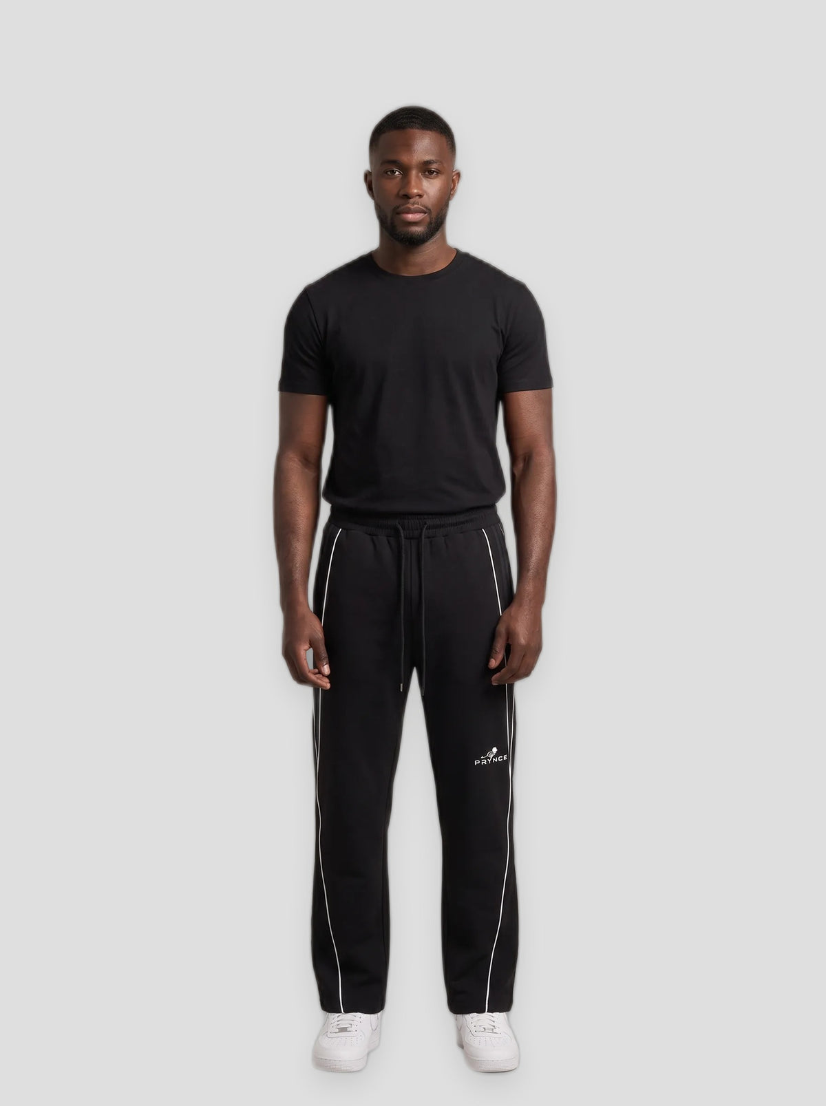 A man modeling a black short-sleeve t-shirt and black sweatpants with white side stripes and a white Adidas logo, standing against a solid grey studio background.