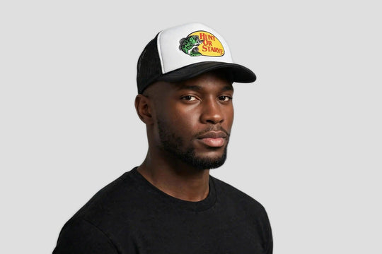 A man wearing a black and white trucker hat with the iconic green and yellow hunt or starve  logo on the front, paired with a black t-shirt against a neutral gray background.