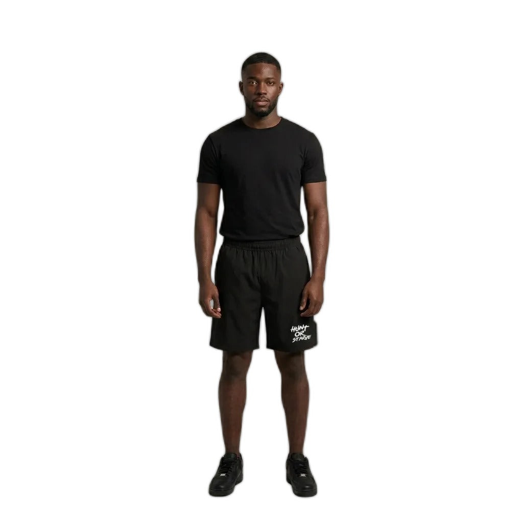 A man stands against a plain light beige studio background wearing a black short-sleeve t-shirt, black athletic shorts with a white logo on the left leg, and black sneakers.