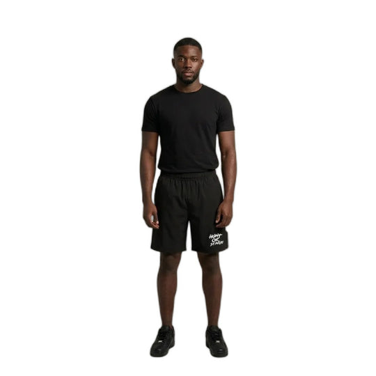 A man stands against a plain light beige studio background wearing a black short-sleeve t-shirt, black athletic shorts with a white logo on the left leg, and black sneakers.