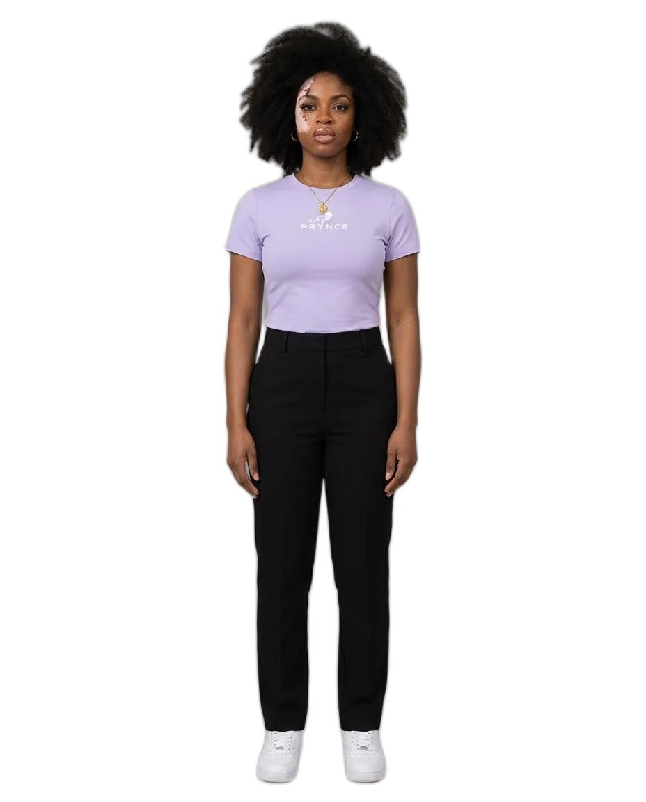 A woman stands against a plain beige studio background wearing a light lavender short-sleeve t-shirt with a small white graphic on the chest, black pants, and white sneakers.