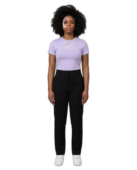 A woman stands against a plain beige studio background wearing a light lavender short-sleeve t-shirt with a small white graphic on the chest, black pants, and white sneakers.