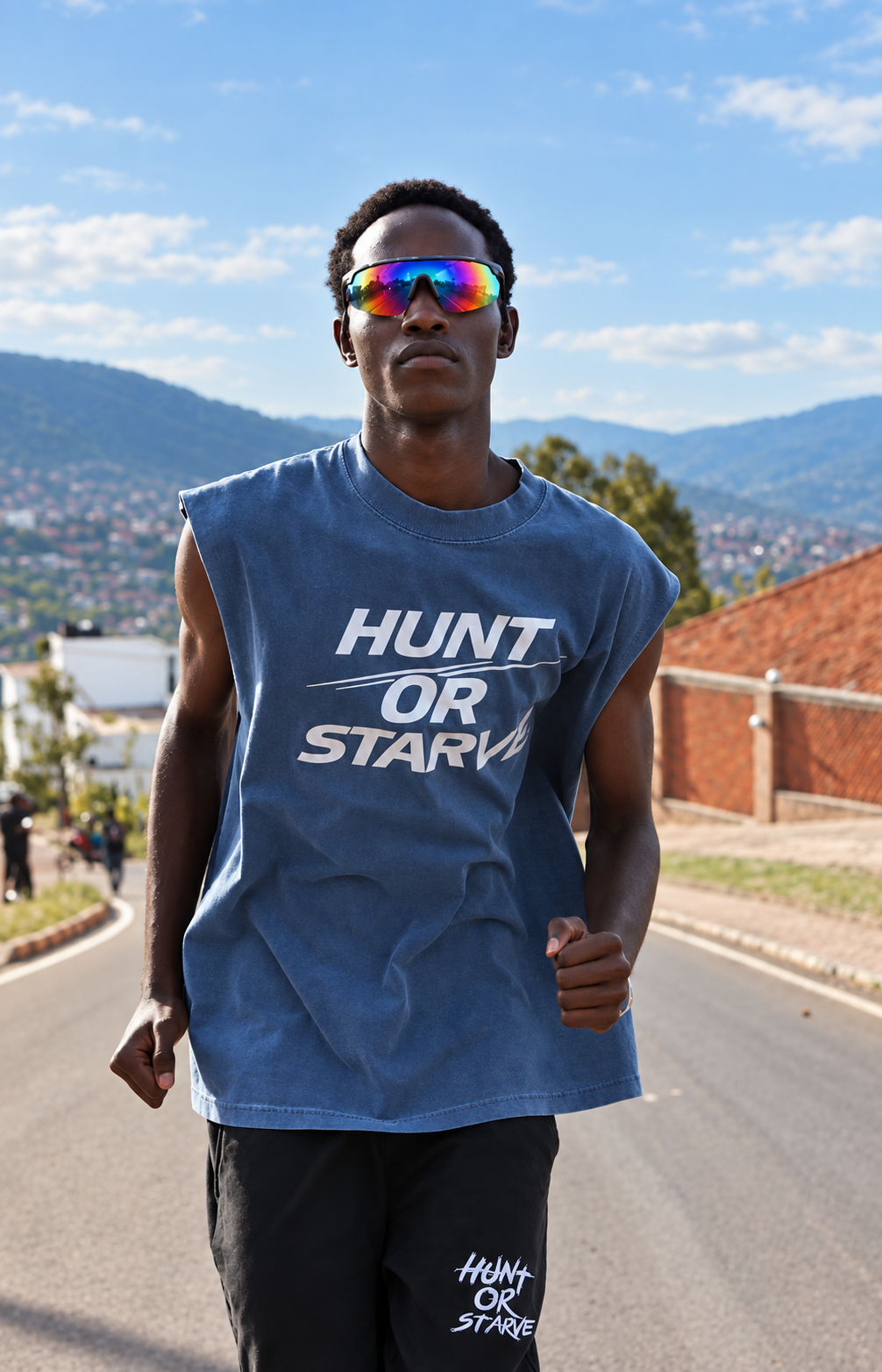 A man wearing a sleeveless blue t-shirt with 'HUNT OR STAY' printed in white on the front, standing on an outdoor road with mountains and a brick wall in the background.