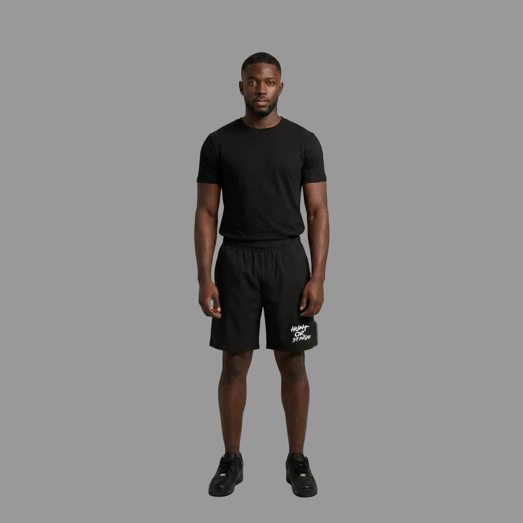 A man stands against a plain light beige studio background wearing a black short-sleeve t-shirt, black athletic shorts with a white logo on the left leg, and black sneakers.