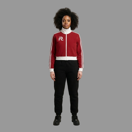A model stands against a neutral beige studio background wearing a red and white track jacket with black sweatpants and black sneakers. The jacket features a white collar, white zipper, white side stripes, and a white &
