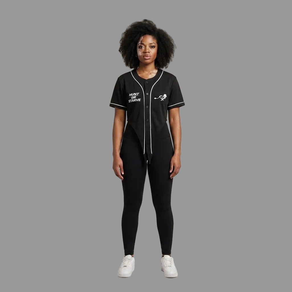 A model stands against a plain beige studio background wearing a black baseball-style jumpsuit with white piping and graphics, black leggings, and white sneakers.