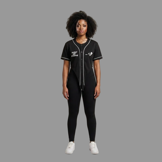 A model stands against a plain beige studio background wearing a black baseball-style jumpsuit with white piping and graphics, black leggings, and white sneakers.