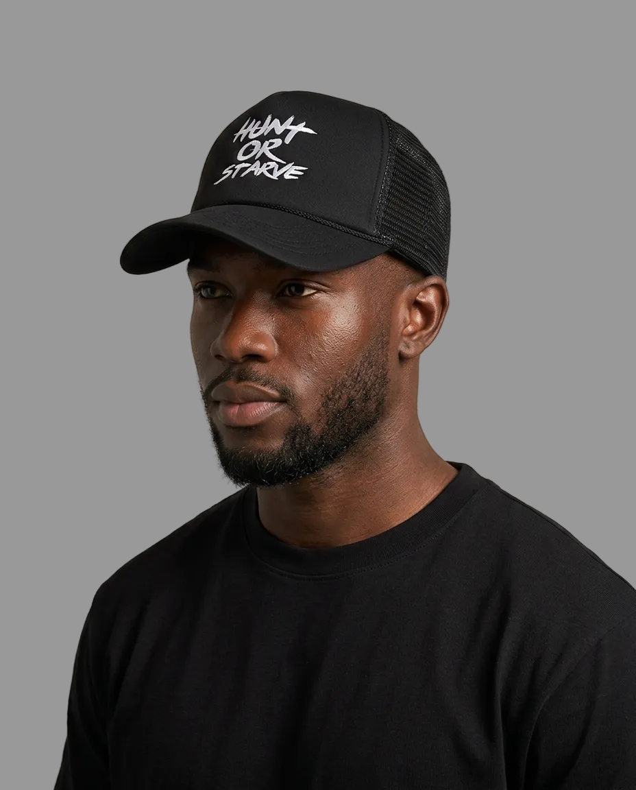 A man wearing a black trucker cap with white embroidered text reading 'HUNT OR STAYE' on the front panel, photographed against a plain light beige studio background.