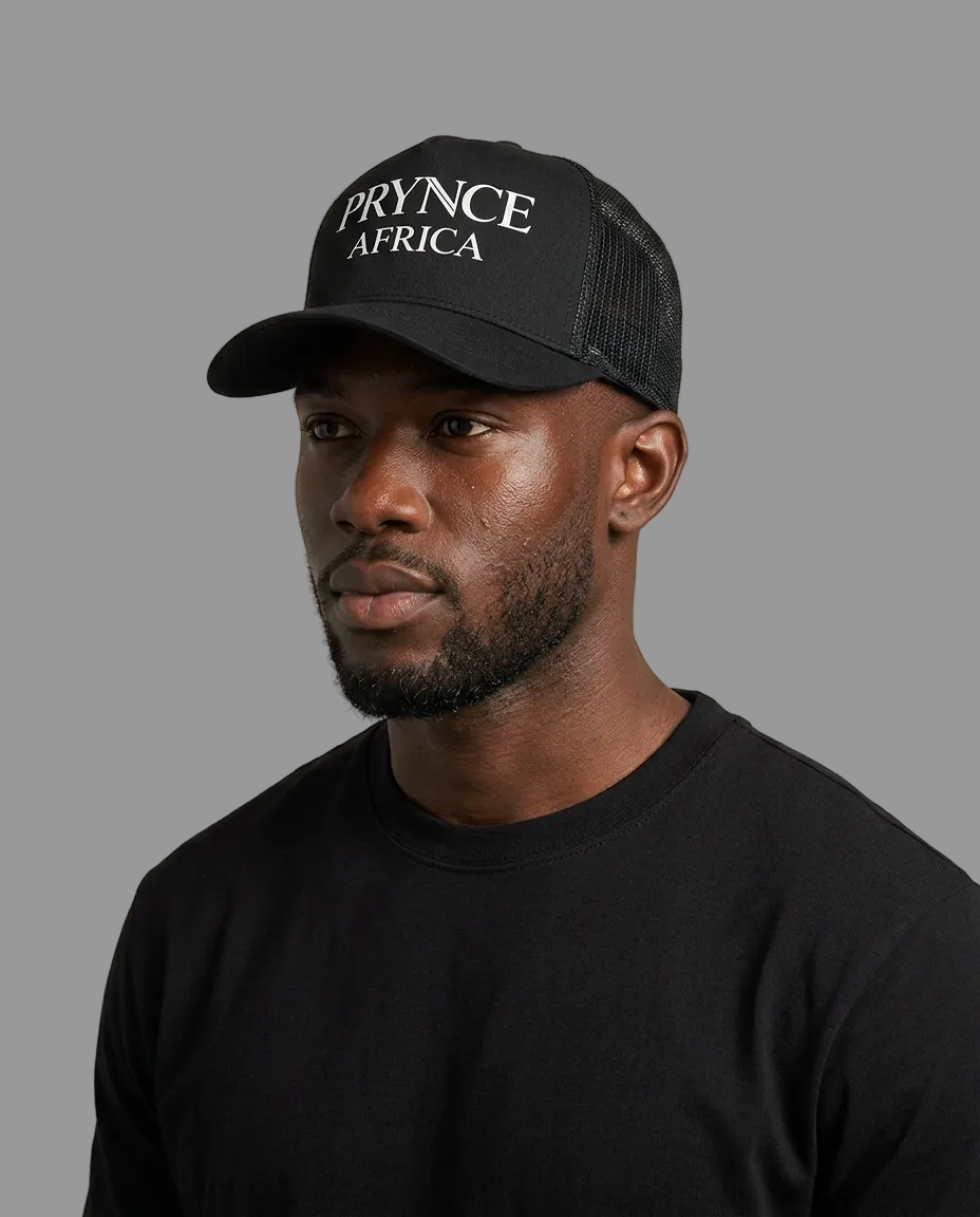 A man wearing a black trucker hat with 'PRYNCE AFRICA' embroidered in white on the front panel, paired with a black t-shirt against a solid light beige background.