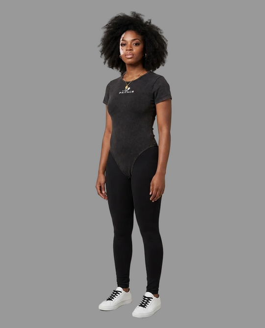 A woman modeling a dark gray short-sleeve bodysuit with a small gold and white logo on the chest, paired with black leggings and white sneakers, against a plain light gray studio background.