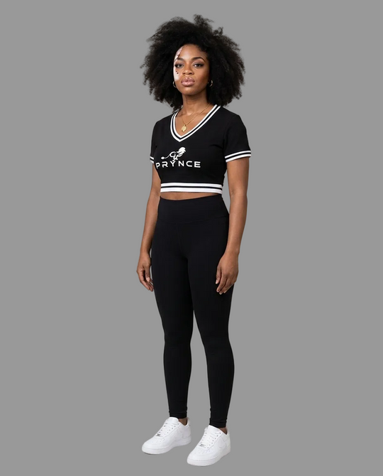A woman modeling a black athletic crop top and matching leggings set with white trim and white sneakers, standing against a plain light beige studio background.
