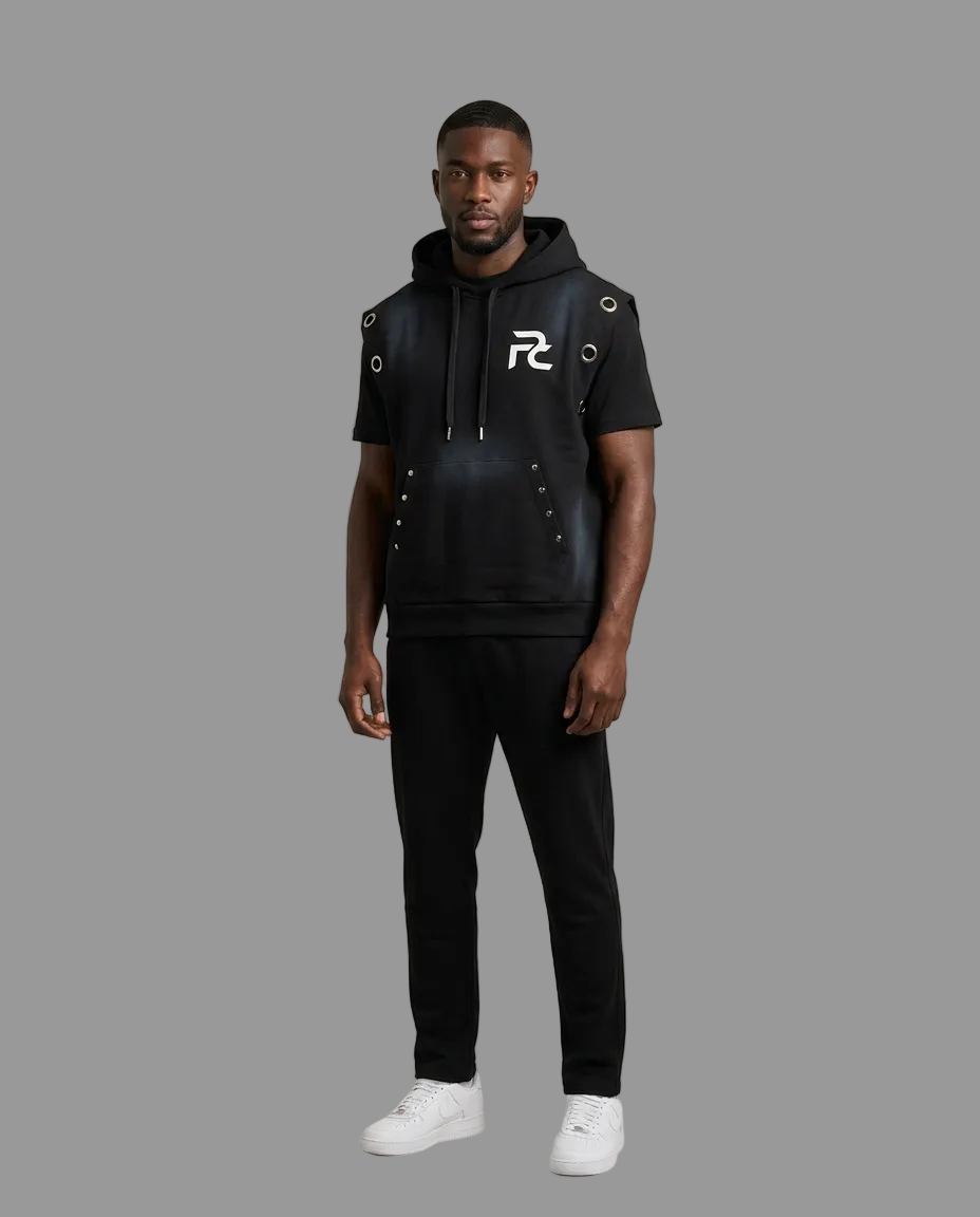 A man modeling a black short-sleeve hoodie with faux-leather paneling, silver grommet details, and a white graphic logo on the chest, paired with black sweatpants and white sneakers, against a solid beige studio background.