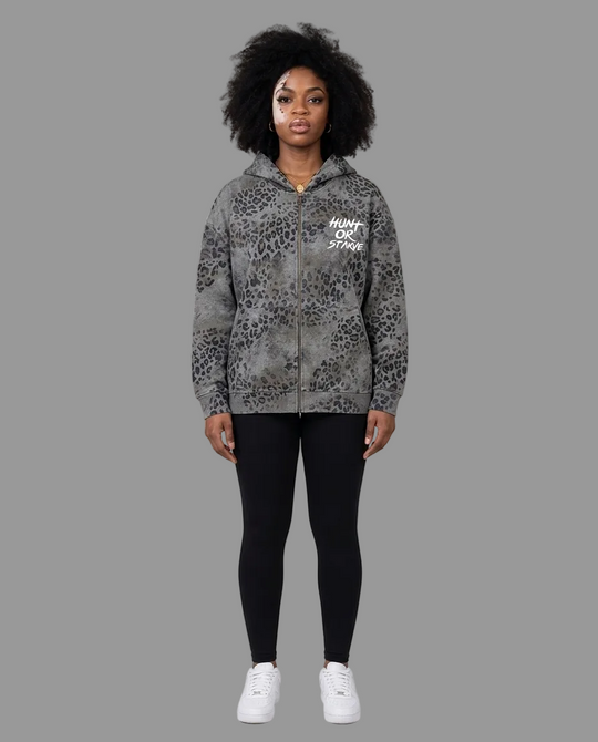 A model stands against a neutral beige studio background wearing a gray and black leopard-print zip-up hoodie with &