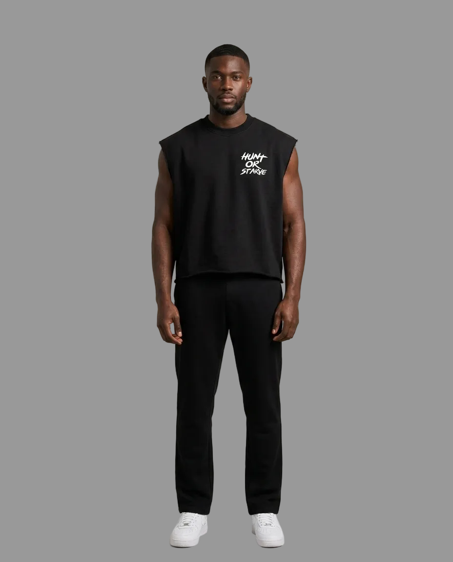 A man modeling a black sleeveless crop top with a white graphic print on the left chest, paired with black pants and white sneakers, against a neutral beige studio background.