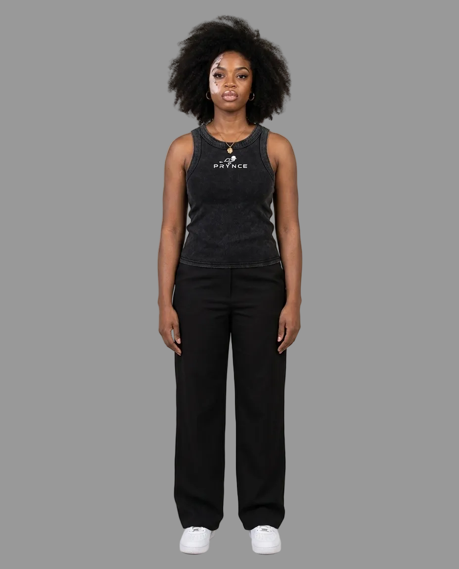 A woman stands against a plain light grey studio background wearing a dark charcoal sleeveless tank top with a white printed logo and black wide-leg pants, paired with white sneakers.