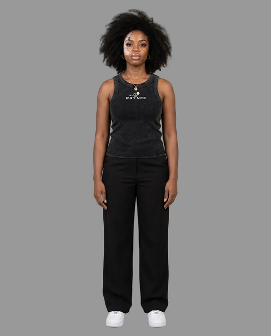 A woman stands against a plain light grey studio background wearing a dark charcoal sleeveless tank top with a white printed logo and black wide-leg pants, paired with white sneakers.