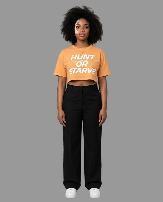 A model stands against a plain beige studio background wearing a cropped orange t-shirt with the text &