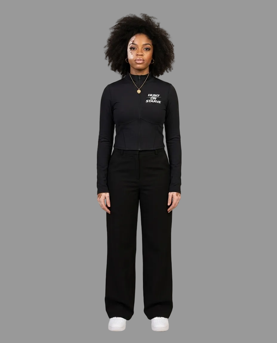 A woman stands against a plain beige studio background wearing a fitted black long-sleeve top with white text on the chest, black wide-leg pants, and white sneakers.