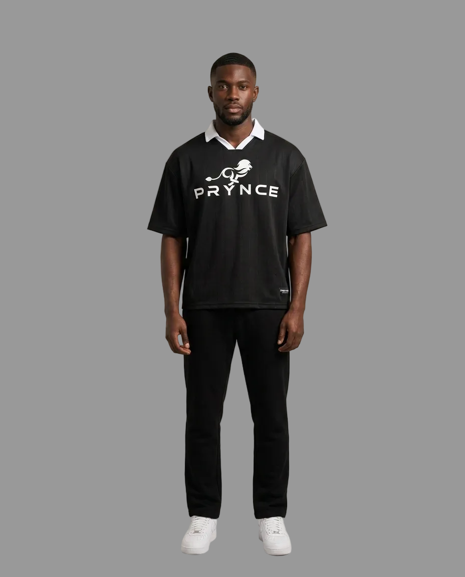 A man modeling a black short-sleeve polo shirt with a white collar, white 'PRYNCE' text and bird logo on the chest, paired with black pants and white sneakers, against a solid light beige studio background.